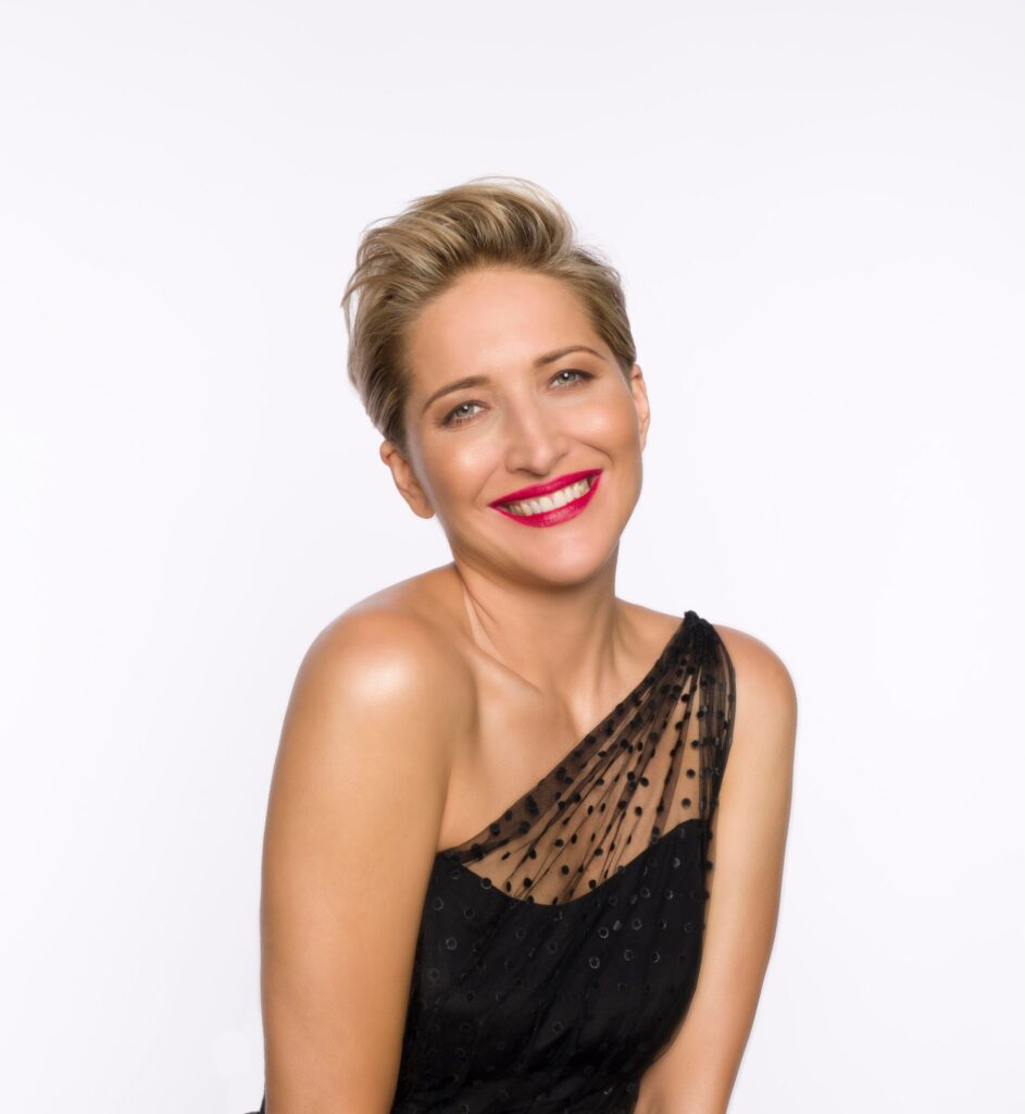 Szeroko uśmiechnięta Maga Steczkowska, ubrana w małej czarnej. Artystka na tle bieli.  Ujęcie do pasa. 