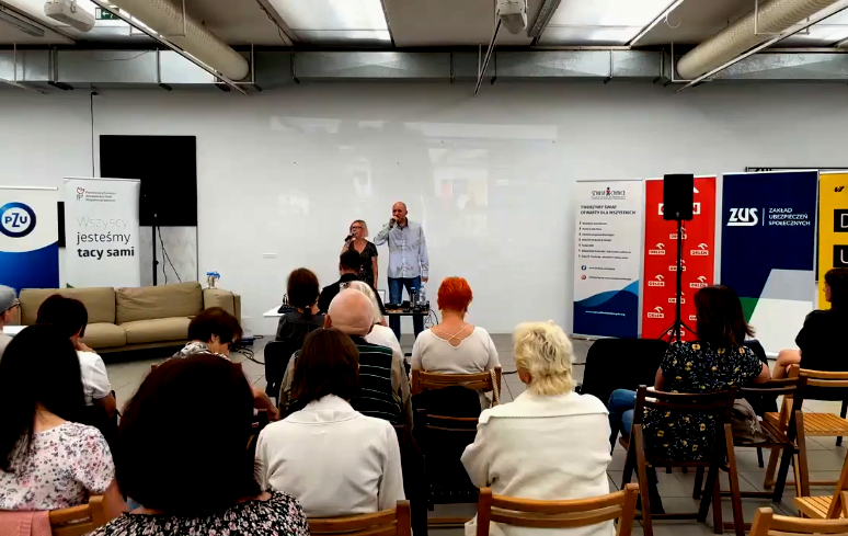 Łukasz i Karolina śpiewają dla publiczności zgromadzonej w sali galerii BWA w Katowicach. Wokół nich banery z logotypami organizatorów i gości zaproszonych do projektu. 