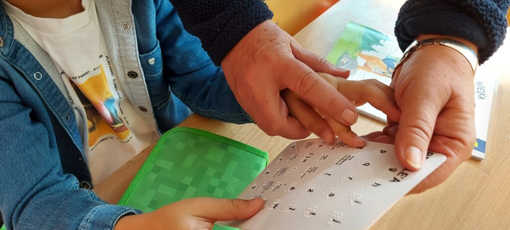 Dłoń dziecka i dłoń nauczycielki na kartce z pismem punktowym 