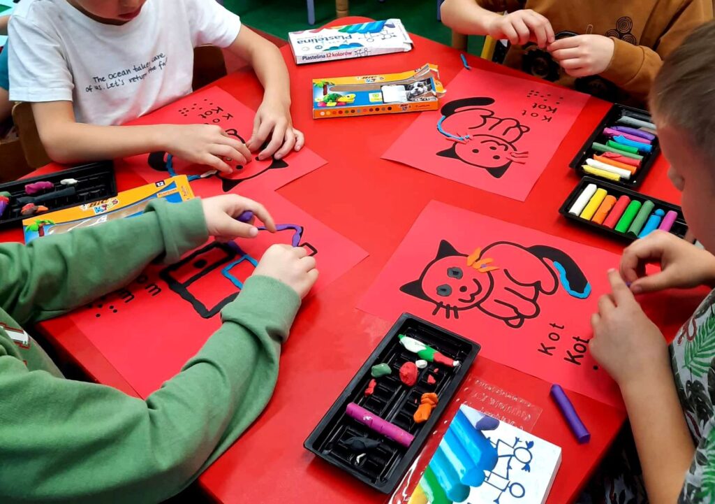 Dłonie dzieci trzymają plastelinę. Wyklejają ilustracje z kotkami i domkami. 