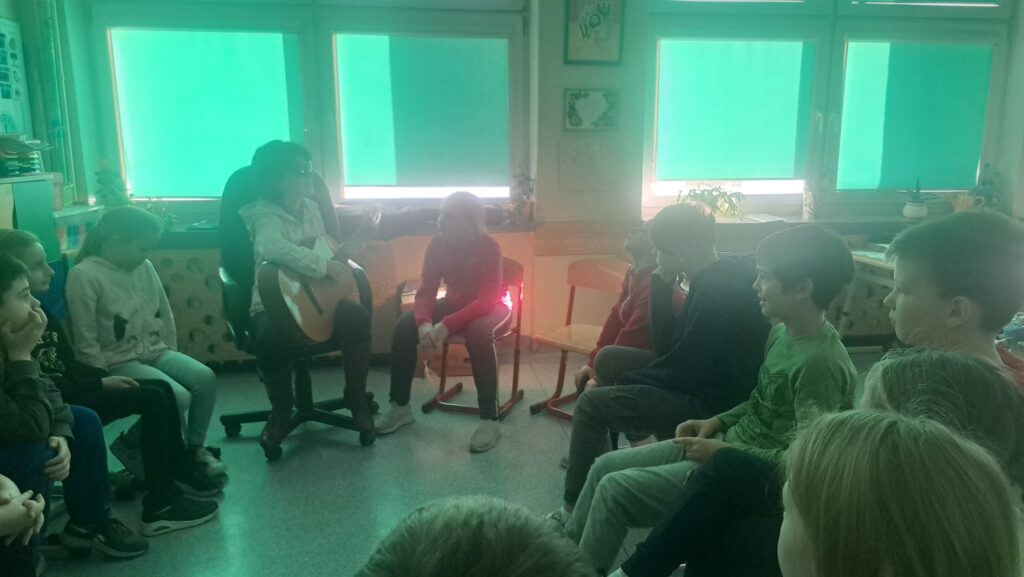 Grupa przedszkolaków i Kasia gra na gitarze. Wszyscy skąpani w lekko zielonym świetle - za sprawą rolet w tej barwie. 
