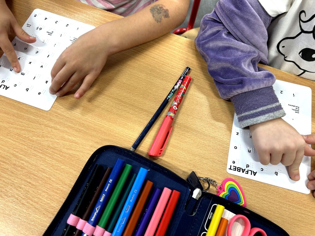 Dziecięce dłonie dotykają wzorników z literami stworzonymi na potrzeby niewidomych. Dla kontrastu - fragment kolorowego piórnika z pisakami i kredkami. Widoczny również na zdjęciu z dołu.
