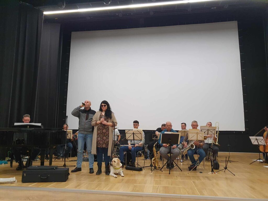 Na scenie: Łukasz, Daria Barszczyk, a obok niej jej pies przewodnik. W oddali - akompaniujący im zespół. 