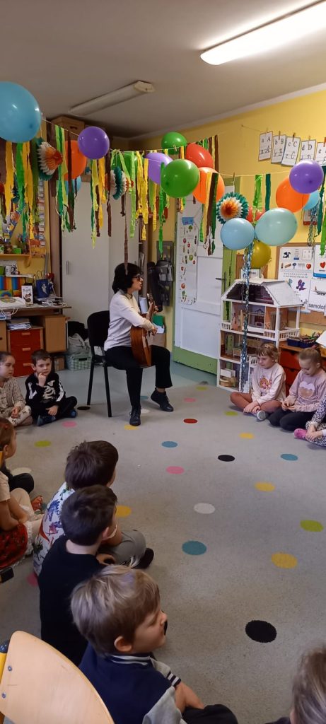 Kolorowa sala z balonami, Kasia gra na gitarze, obok niej zasłuchane przedszkolaki. 