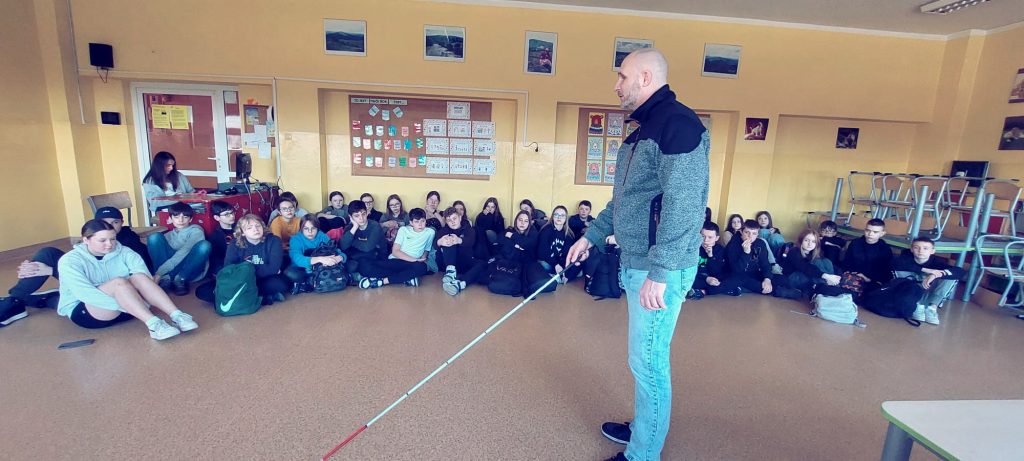 Łukasz Baruch porusza się po świetlicy z białą laską, obserwują go uczennice i uczniowie.