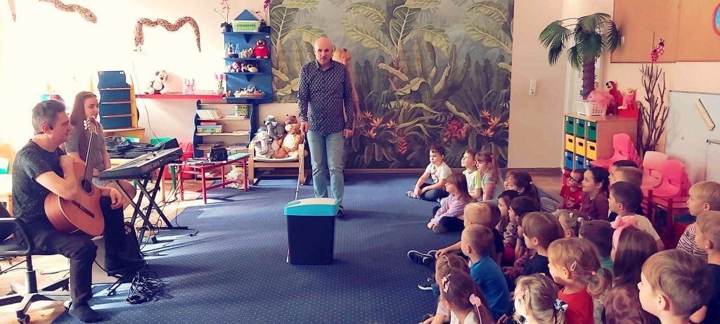 Łukasz pokazuje dzieciom, jak omija przeszkody dzięki białej lasce. na fotografii: Żenia z gitara, Rita przy instrumencie klawiszowym i dziecięca publiczność.