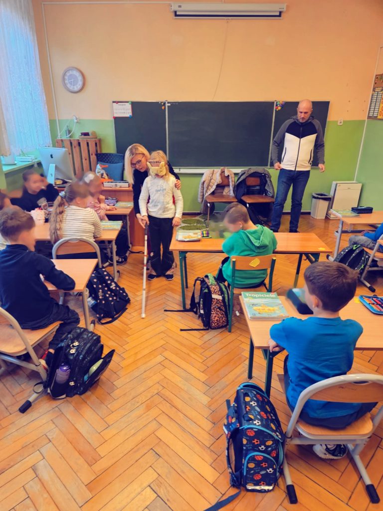 Łukasz Baruch i Karolina Żelichowska oraz dzieci w klasie. Karolina pomaga uczestniczce w zadaniu na orientację po sali, która ma zasłonięte oczy opaską i używa białej laski.