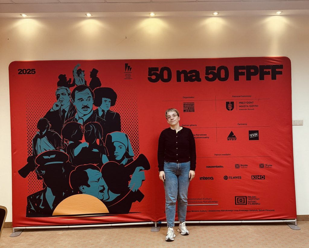 Justyna na tle czerwonego baneru z napisem 50 na 50 FPFF. Obok Justyny narysowane postaci bohaterów takich jak: Ida, Hans Kloss, czy amator z kamerą z filmu K. Kutza. Na banerze logotypy - w tym Polskiego Instytutu Sztuki Filmowej, Ministra Kultury, stacji telewizyjnych, portali www i innych organizacji.