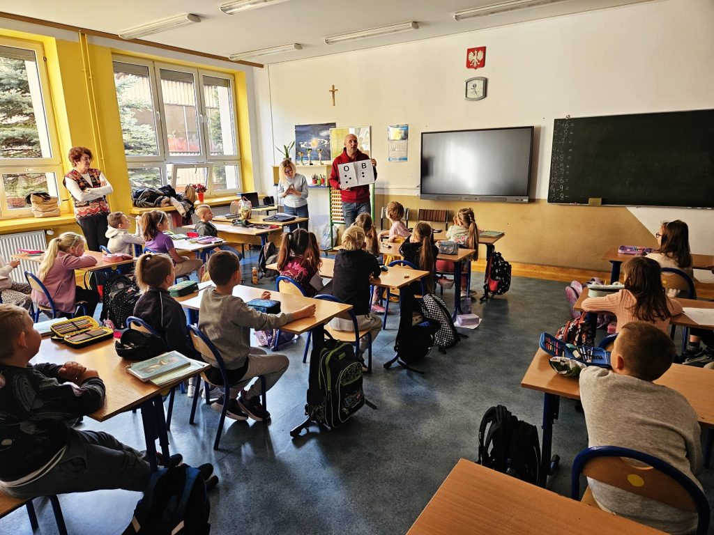 Łukasz trzyma wydrukowany wzór z literami O i B, a Karolina przy biurku, trzyma w dłoniach wzorniki z pismem punktowym, które zaraz rozda dzieciom, siedzącym w ławkach.