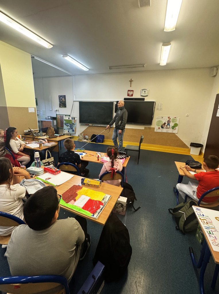 Łukasz Baruch w sali. Dzieci w ławkach. Obserwują jak używa białej laski. 