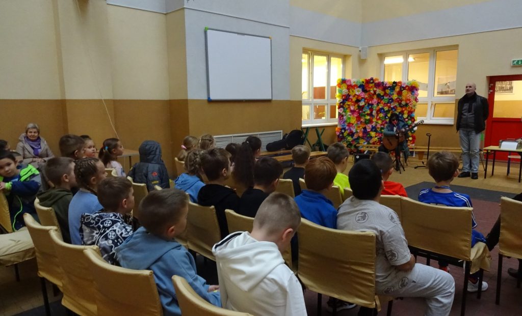 Spotkanie w ZSP7 - dzieci w holu siedzą na krzesełkach, Kasia gra na gitarze, obok niej Łukasz Baruch. 