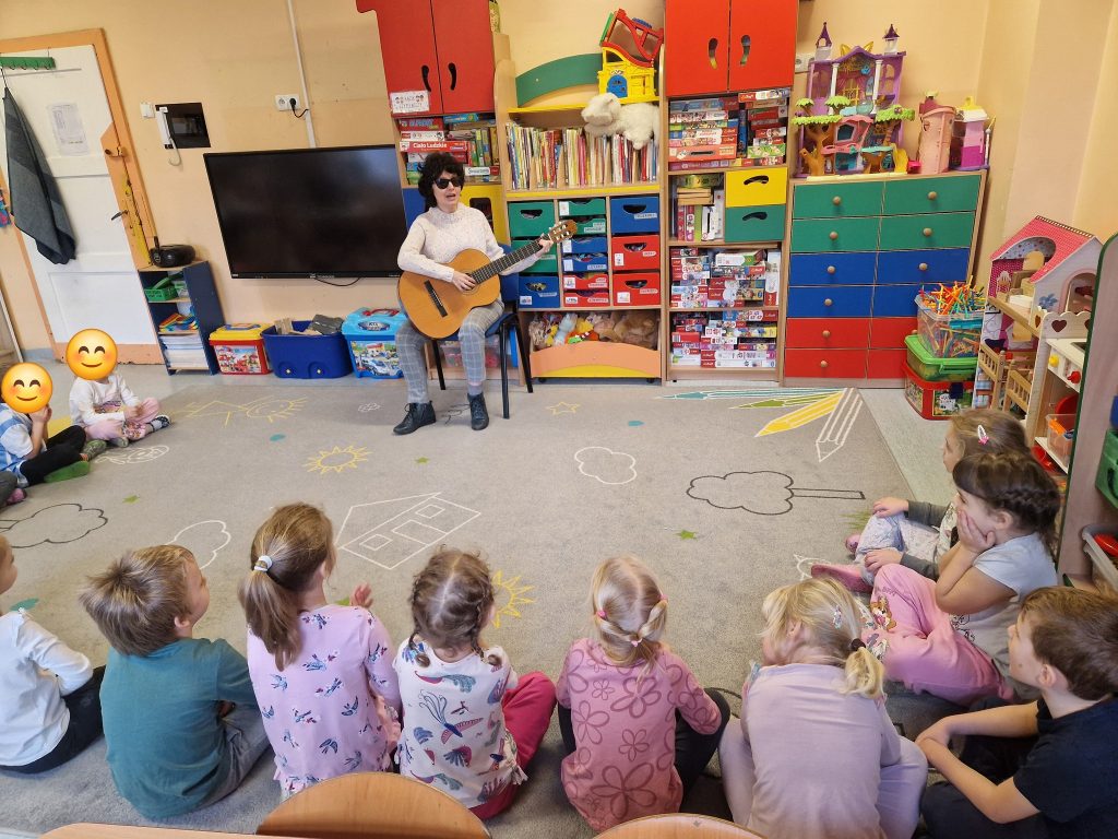 Na fotografii: Kasia i dzieci. Ona gra na gitarze.  Dzieci patrzące na wprost, mają kolorowy stempelek - anonimowa buzia. 