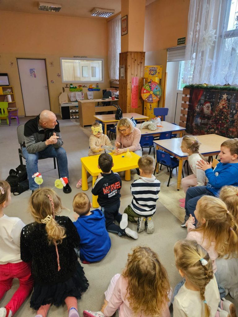 Dziecko ma zasłonięte oczy opaską, nalewa wody do kubeczka. W zadaniu  pomaga mu Karolina. Inne przedszkolaki obserwują. Łukasz Baruch nasłuchuje, co się dzieje. 