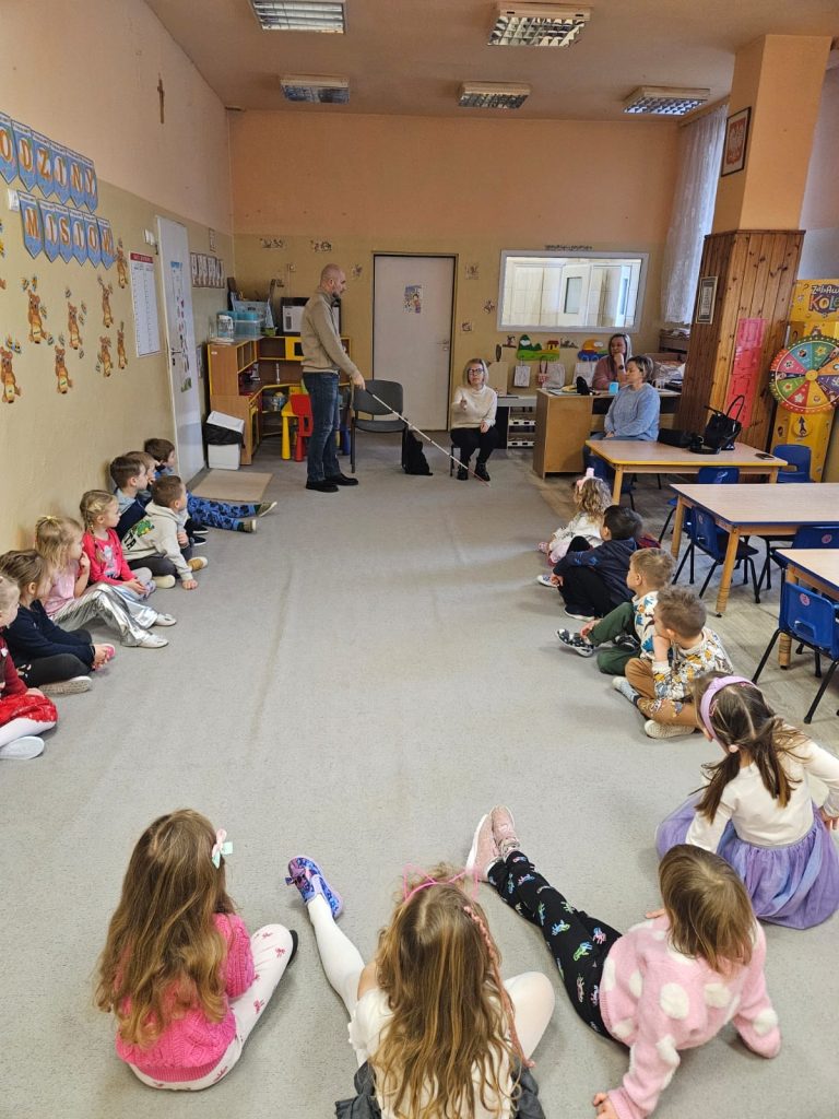 Łukasz Baruch z białą laską, dzieci w półkolu. Obok niego Karolina Żelichowska. 