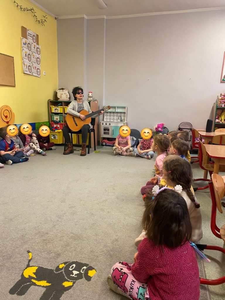 Kasia w towarzystwie przedszkolaków. Gra na gitarze, dzieci w kręgu. Ich twarze pozasłaniane wesołymi minkami. 