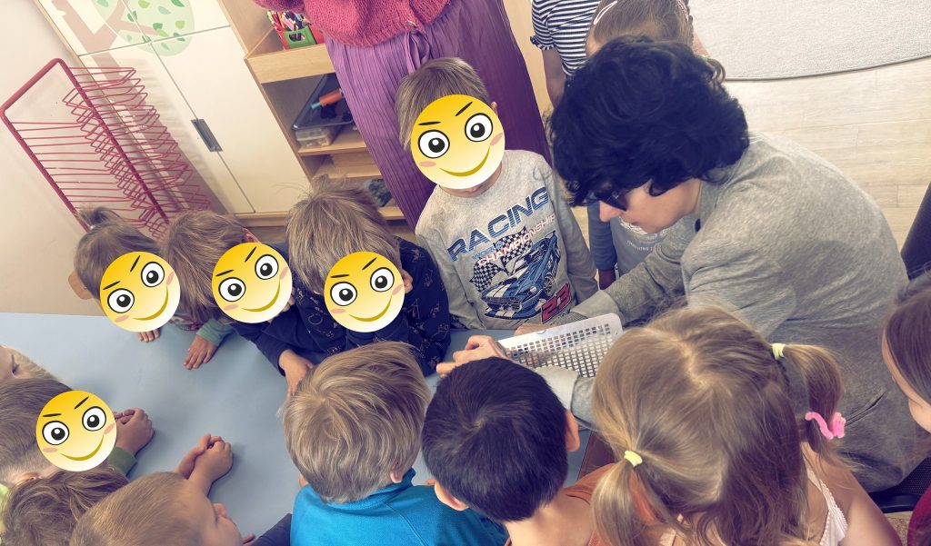 Kasia pokazuje dzieciom do czego służy tabliczka brajlowska i dłutko. Wokół niej przedszkolaki - niektóre mają naklejki - minki w miejscach twarzy.