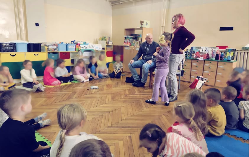 Łukasz Baruch i Karolina Żelichowska oraz uczestnicy warsztatów. Jedno z dzieci ma zasłonięte oczy opaską i próbuje odszukać maskotkę.
