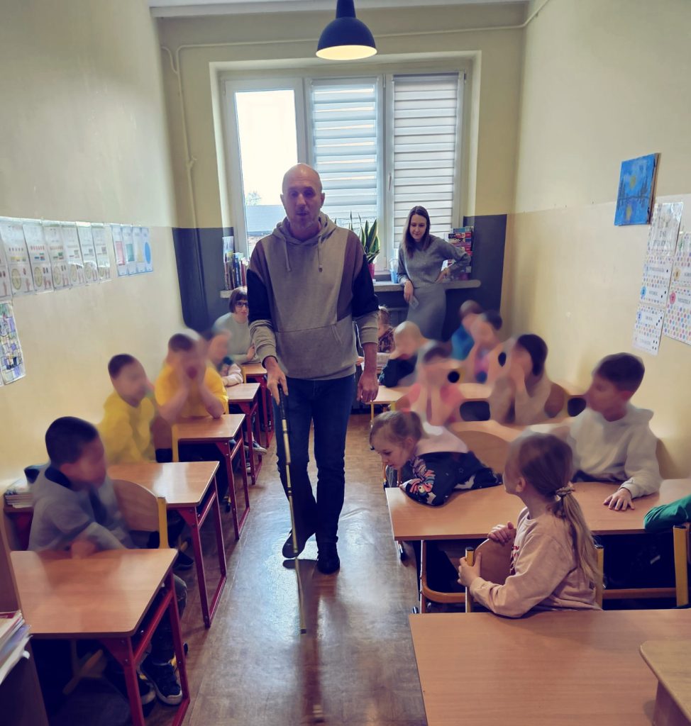 Projekt IPE MOON - Łukasz Baruch w sali lekcyjnej, dzieci w ławkach. Obserwują, jak niewidomy używa białej laski do orientacji w ciasnej przestrzeni sali.