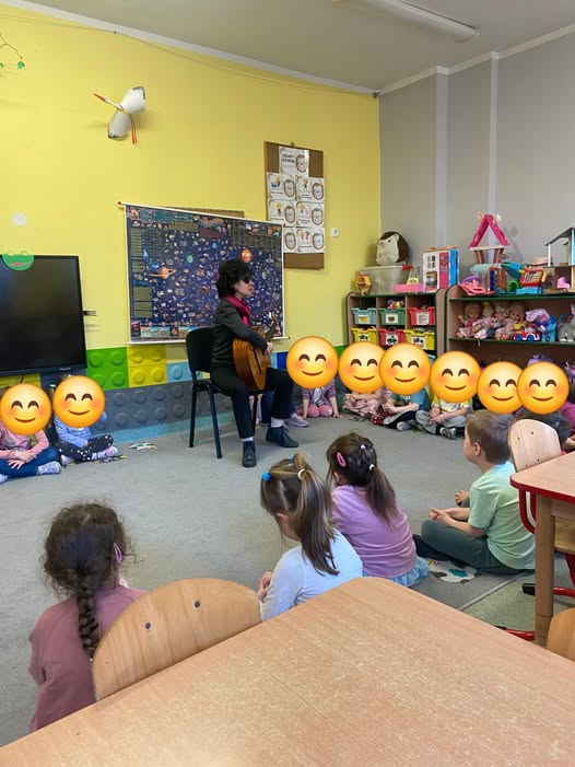 Katarzyna Nowak gra na gitarze, wokół niej dzieci - część z nich ma buzie zasłonięte kolorowymi naklejkami. 