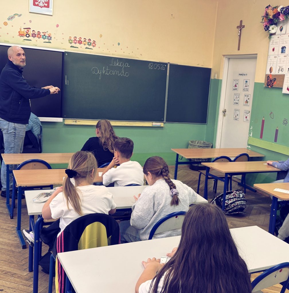 Uczestnicy zajęć, dotykają wzorników z pismem punktowym, a Łukasz opowiada o pisaniu na maszynie brajlowskiej, wykonując gest jakby dotykał klawiszy. Na szkolnej tablicy widnieje napis DYKTANDO. 😉
