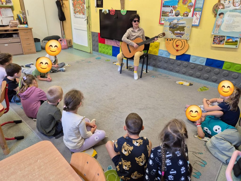 Warsztaty muzyczne - Katarzyna Nowak gra na gitarze. Inna grupa dzieci. Maluchy mają pozaklejane buzie na fotografii - emotkami.