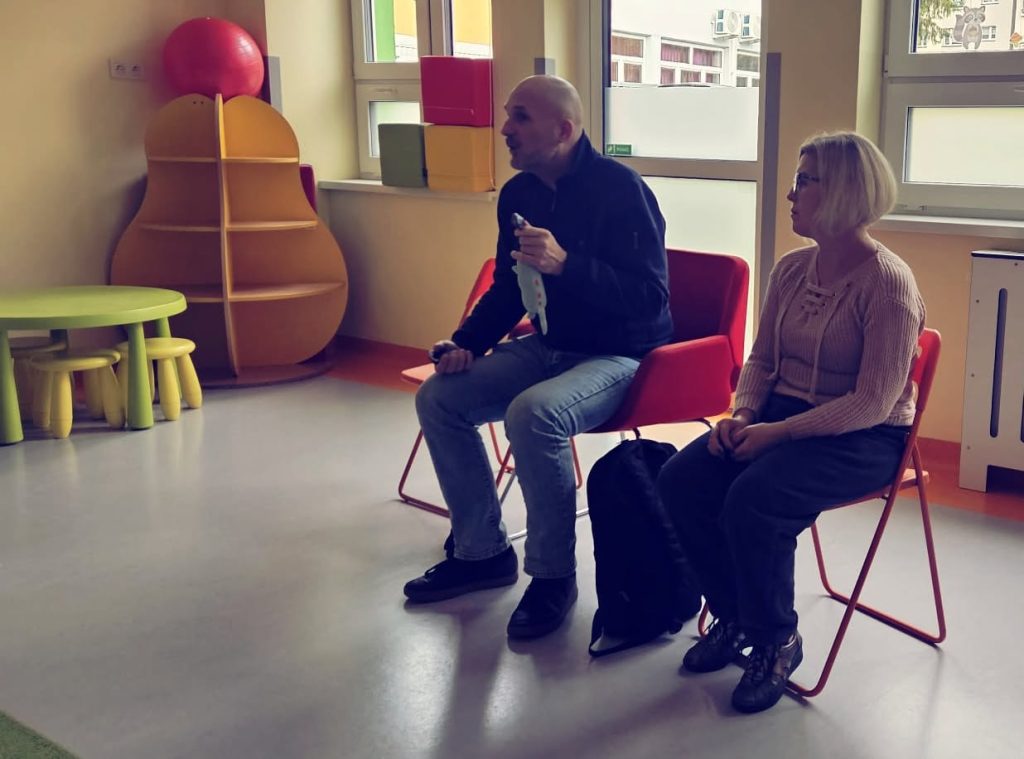 Łukasz Baruch i Karolina Żelichowska pokazują maskotkę – kotka. Siedzą na krzeskach w sali przedszkolnej. 