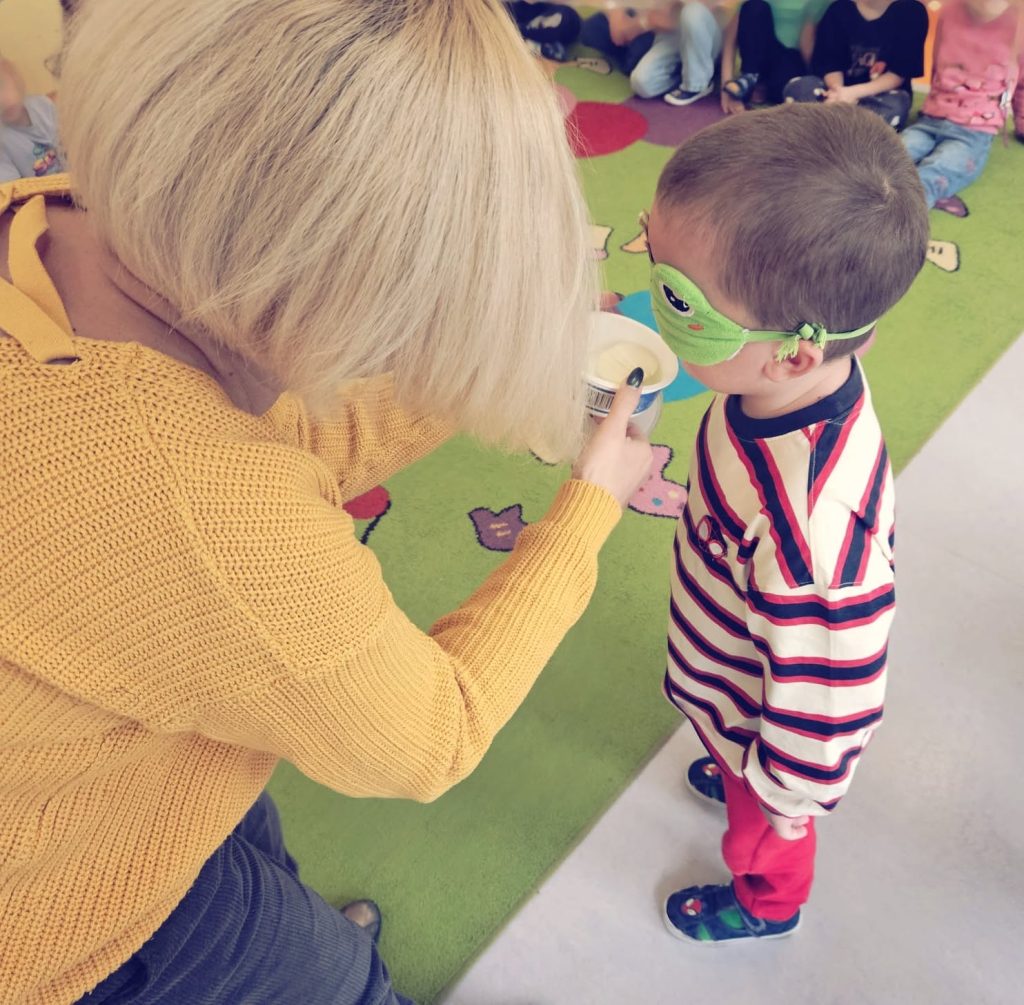 Koralina podaje pudełeczko. Dziecko ma zasłonięte opaską oczy, wącha, co jest w środku.